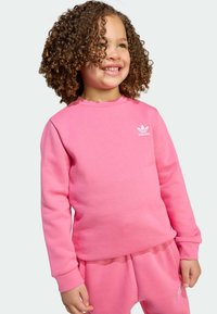 Ružová mikina vyrobená z mäkkého materiálu, s okrúhlym výstrihom a dlhými rukávmi, so malým bielym logom Adidas na hrudi.