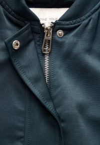 Ciemnozielona kurtka bomber z gładką fakturą, srebrny zamek błyskawiczny oraz zatrzaskowy detal przy kołnierzu. Widoczna wewnętrzna metka.