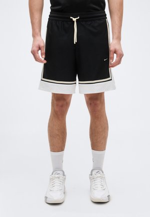 Mężczyzna ubrany w czarno-białe krótkie spodenki do koszykówki Nike, białe skarpetki i białe sportowe buty, stojący na jednolitym tle.