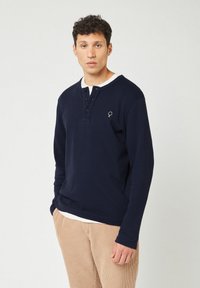 Maglietta henley a maniche lunghe blu navy con colletto bianco, dotata di tre bottoni e un piccolo logo ricamato sul lato sinistro del petto. Texture a nido d'ape.