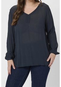 Blouse à manches longues bleu marine foncé avec un col en V, tissu texturé, coupe ample et poignets retroussés, associée à un jean bleu foncé.