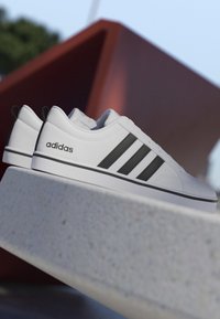 Zapatillas blancas Adidas con rayas negras apoyadas en un borde de hormigón contra un fondo exterior desenfocado con una estructura geométrica roja.