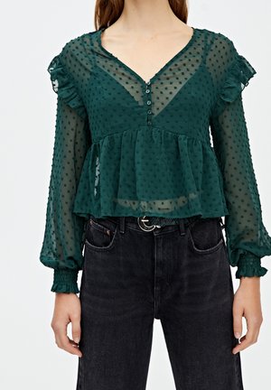 Kvinde iført en gennemsigtig mørkegrøn bluse med teksturerede prikker, flæseærmer, knappet V-hals og sorte højttaljede jeans med bælte.