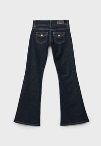 Tmavomodré riflové džínsy s rozšíreným strihom nohavíc, dvoma zadnými vreckami s gombíkmi a kontrastným prešívaním na jednoduchom svetlom pozadí.