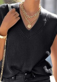 Žena v černém žebrovaném topu bez rukávů s V-výstřihem, s vrstvenými zlatými řetízky a černými kalhotami s vysokým pasem, drží popruh od kabelky.