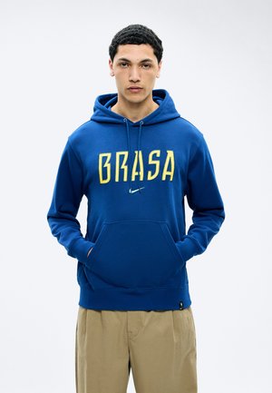 Young man with short curly hair wearing a blue hoodie with yellow "BRASA" text and beige pants, hands in front pocket, neutral background.