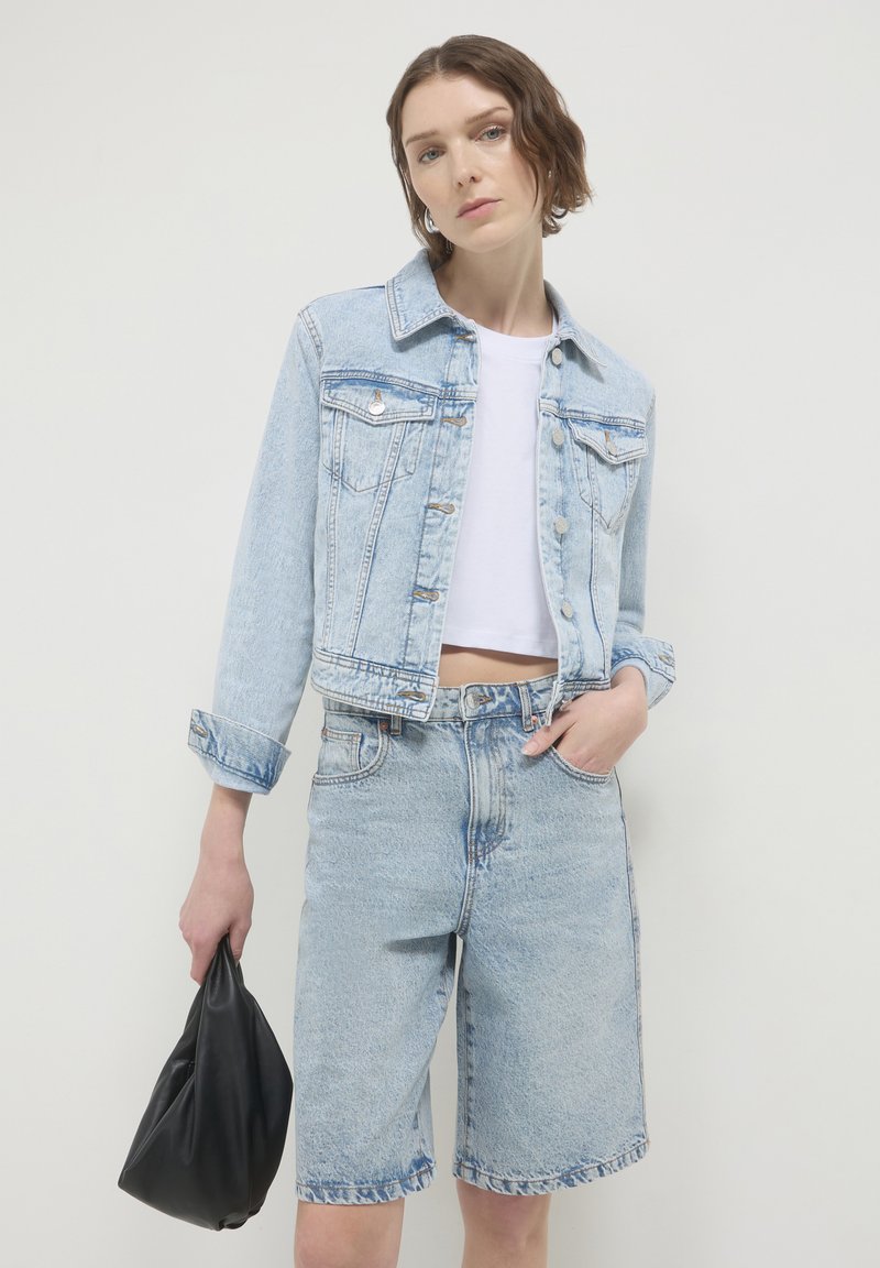 Donna che indossa una giacca di denim chiaro e pantaloni shorts con un crop top bianco, mentre tiene una borsa nera morbida contro uno sfondo bianco semplice.