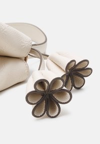 Krämfärgad handväska med en texturerad yta, som har blommformade ändar. Detaljer inkluderar bruna accenter och släta, rundade former.