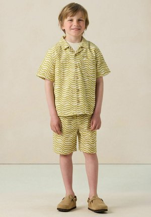 Garçon souriant debout, portant une chemise à manches courtes à motifs ondulés jaunes et blancs et un short assorti avec des chaussures slip-on beige.