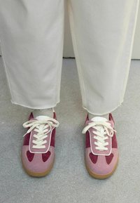 Rosa och vinröda sneakers med en texturerad kordflorsöverdel, vita snören och en gummisula, kombinerade med krämfärgade byxor.