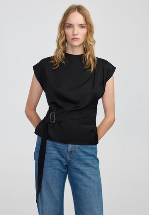 Vrouw met golvend blond haar, gekleed in een zwarte mouwloze top met een zijgespeldetail en blauwe jeans, staande tegen een effen lichte achtergrond.