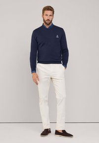 Maglione navy indossato sopra una camicia blu chiaro, con pantaloni crema e scarpe slip-on marrone scuro. Presenta un piccolo logo sul maglione.