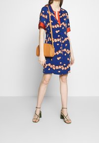 Bloemige marineblauwe jurk met rode accenten, korte mouwen en een strik taille. Accessoires zijn een tan schoudertas en groene gespenhakken.