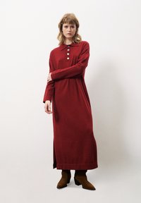 Robe rouge à manches longues avec un col, des détails de boutons et une fente sur le côté, fabriquée en tissu doux. Associée à des bottes chelsea à talons marron.