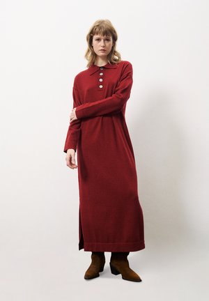 Langsleeve rode jurk met een kraag, knoopdetails en een zijspleet, gemaakt van een zachte stof. Gecombineerd met bruine enkellaarsjes met hak.