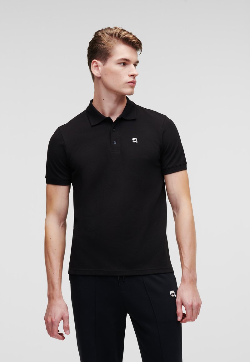 Polo shirt nera in cotone, con colletto, maniche corte e un piccolo logo bianco sul petto. Abbinato a pantaloni neri.