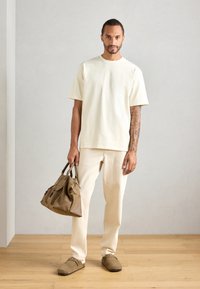 Uomo in t-shirt beige e pantaloni sta in piedi su un pavimento in legno, tenendo una borsa in pelle marrone nella mano destra, davanti a una parete liscia grigio chiaro.