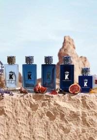Cinq flacons de Cologne Dolce & Gabbana K avec bouchons couronne exposés sur une surface en pierre avec des figues et des fleurs violettes.