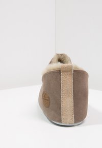 Brown suede slipper with a cream fur lining, prominent back seam, and a circular leather logo detail on the side. Soft texture and rounded shape.