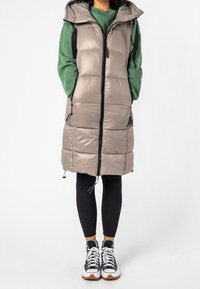 Osoba ubrana w długi beżowy kamizelkę puchową z kapturem, pod której znajduje się zielona bluza, czarne leginsy prążkowane i czarno-białe sneakersy.