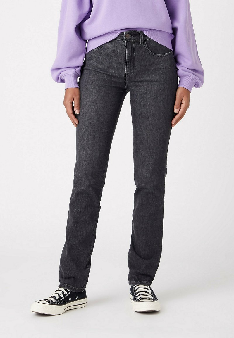 Personne portant un jean droit gris foncé taille haute, un sweat lavande et des baskets basses noires, debout devant un fond blanc.