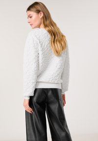 Weißer strukturierter Sweatshirt mit Rundhalsausschnitt und gerippten Bündchen, kombiniert mit schwarzen, weiten Ledehosen. Glattes Haar fällt über die Schulter.