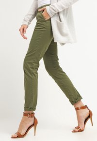 Olive green tailored pants with side pockets, cuffed hems, and a smooth fabric finish, paired with brown high-heeled sandals.