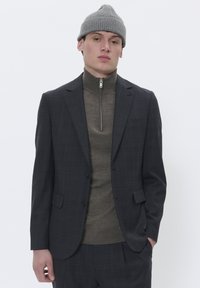 Mand iført en grå strikket beanie, mørkternet blazer og grå lynlås-sweater står mod en ensfarvet hvid baggrund.