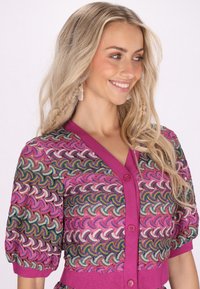 Färgglad stickad cardigan med rosa bas, vågmönster i blått, grönt och gult, korta puffärmar och V-ringning med knappar.
