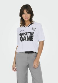 Biała koszulka sportowa z czarnym napisem "RAISE THE GAME", z czarnym kołnierzykiem w serek, w połączeniu z jasnoszarymi spodniami.