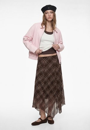Młoda kobieta w czarnym berecie, jasnoróżowej kurtce, białej i brązowej warstwowej bluzce, brązowej asymetrycznej spódnicy w wzory oraz brązowych balerinach.