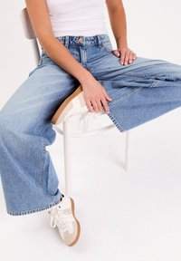 Jean large taille haute bleu clair avec une coupe ample, associés à des baskets blanches avec des accents beige. Tenue décontractée sur une chaise.