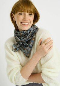 Pañuelo de seda con diseño cachemira azul y marrón atado alrededor del cuello, combinado con un suéter de punto texturizado color crema. Anillo en una mano, fondo neutro.