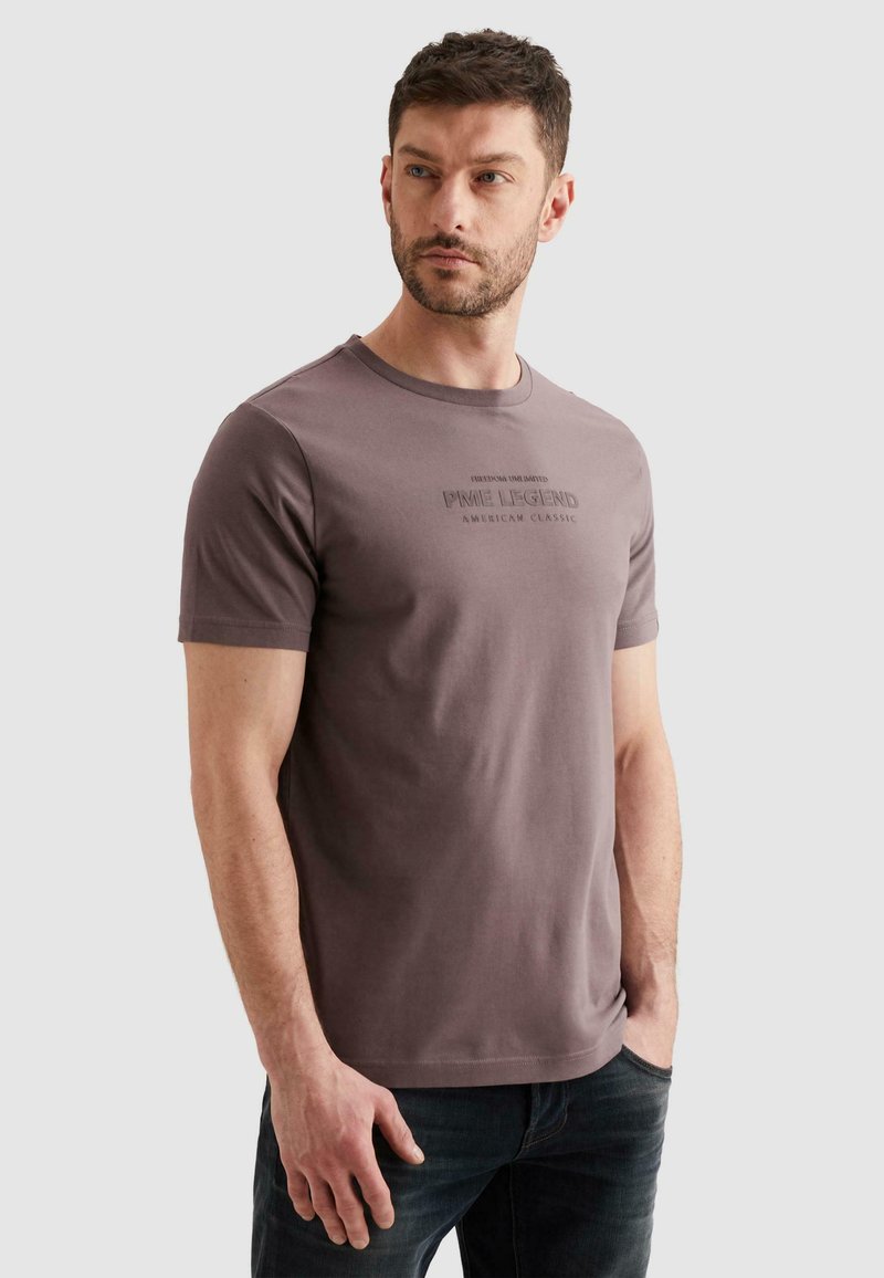 Homme aux cheveux courts portant un t-shirt mauve à manches courtes avec le texte "PME Legend American Classic", debout les mains dans les poches.