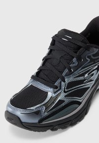 Juodas ir pilkas Skechers sportinis batelis su tinklelio audiniu, sintetinių medžiagų detalėmis ir raišteliais, įstrižai rodomas, kad būtų matyti priekiniai ir šoniniai detalės.