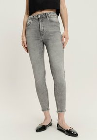 Kobieta w jasnoszarych, wysokich, dopasowanych dżinsach, czarnej krótkiej bluzce i czarnych lakierowanych balerinach stoi na białej podłodze.