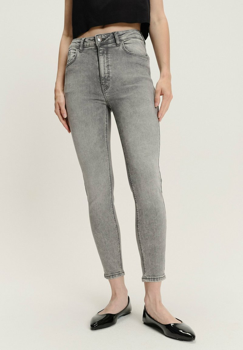 Kobieta w jasnoszarych, wysokich, dopasowanych dżinsach, czarnej krótkiej bluzce i czarnych lakierowanych balerinach stoi na białej podłodze.