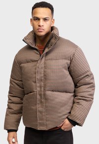 Doudoune à carreaux marron et beige avec un col haut, dotée d'une fermeture éclair et de détails de couture visibles, portée sur un pull sombre.