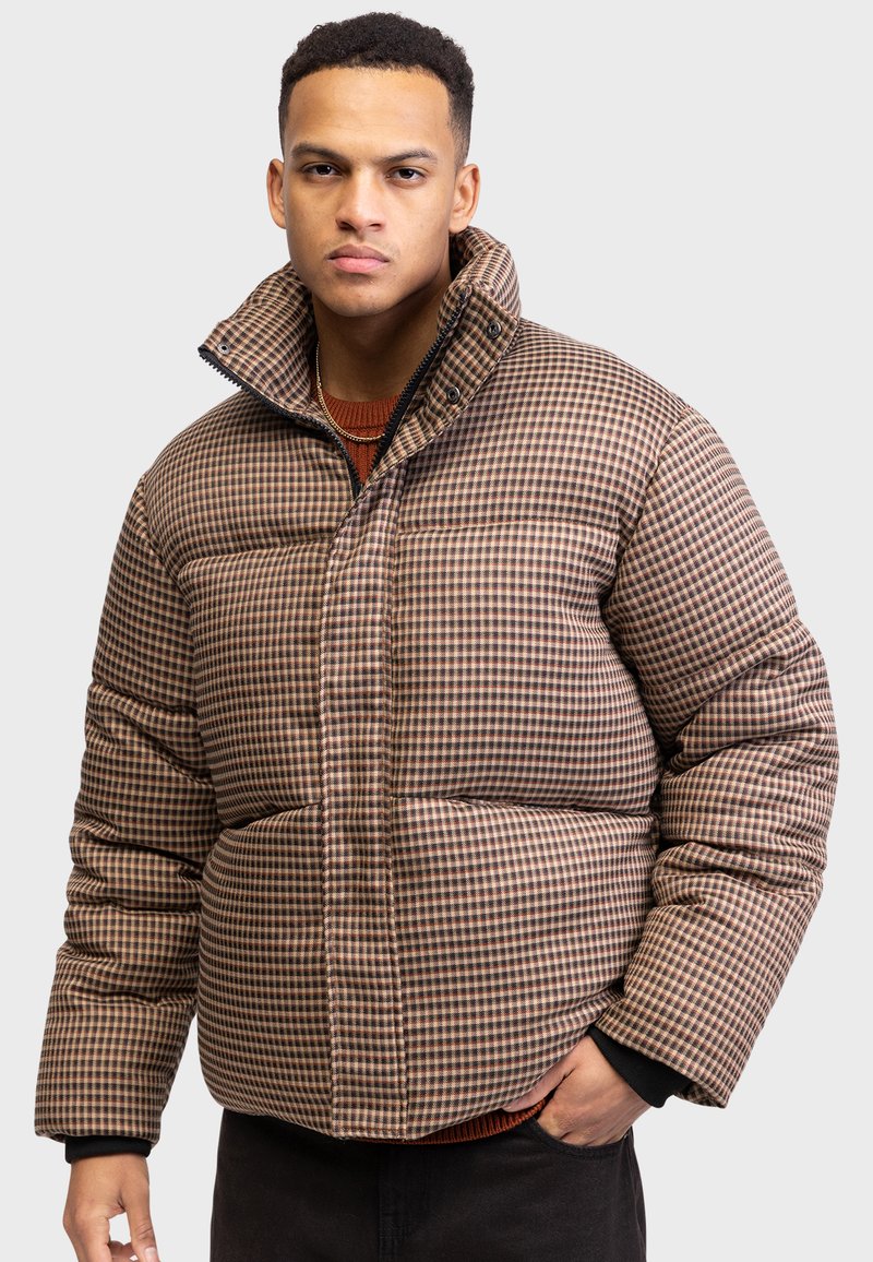 Doudoune à carreaux marron et beige avec un col haut, dotée d'une fermeture éclair et de détails de couture visibles, portée sur un pull sombre.