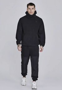 Svart hoodie med hög krage, kombinerad med matchande svarta joggingbyxor. Båda har en slät textur och minimalistisk design.