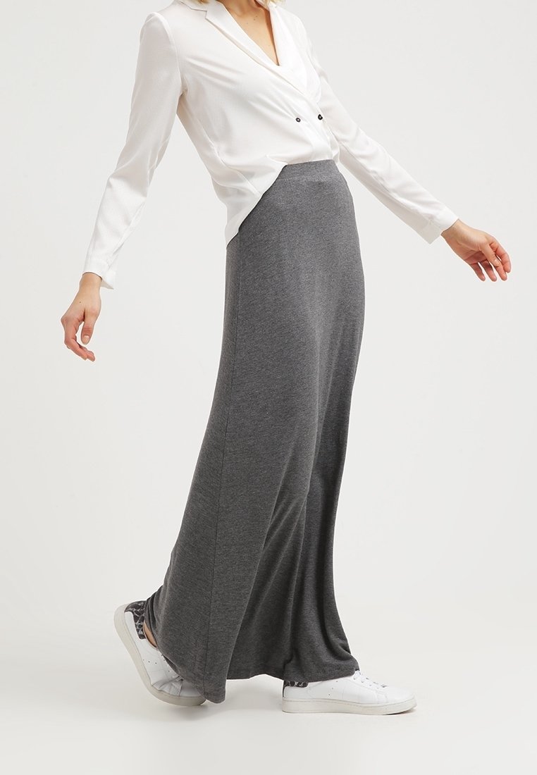 Femme portant un chemisier blanc, une longue jupe grise et des baskets blanches, debout avec les bras légèrement écartés sur fond uni.