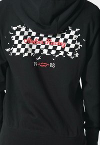 Felpa nera con cappuccio che presenta una grafica di bandiera a scacchi in bianco e rosso sul retro, con il testo "Atelier Racing" e ulteriori dettagli in basso.