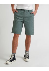 Muž v zelených šortkách do kolen, černých vysokých teniskách, šedých ponožkách a bílém tričku, stojící s rukama v kapsách.