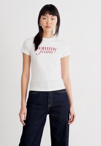 Balta medvilnė marškinėliai su trumpomis rankovėmis, su raudonu "tommy jeans" tekstu. Dėvimi su tamsiai mėlynomis džinsinėmis kelnėmis. Paprasta, prigludusi dizaino forma.