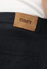 Ετικέτα υφάσματος με την επιγραφή "OBEY" στη μέση μαύρου παντελονιού, φορεμένου με λευκό ριπέ πουκάμισο.