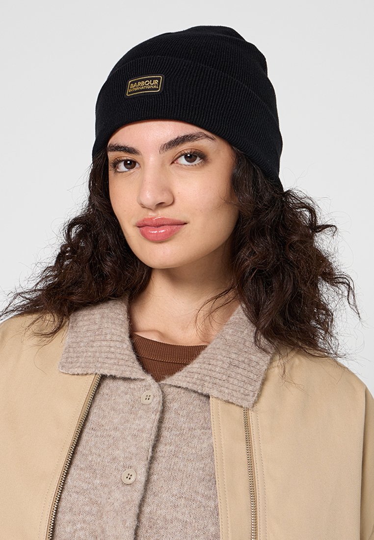 Czarna, prążkowana czapka beanie z małą, złotą prostokątną aplikacją, noszona z beżową kurtką z kołnierzykiem i jasnobrązowym swetrem pod spodem.