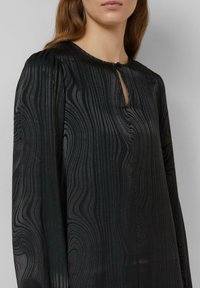 Schwarze Bluse mit langen Ärmeln und welliger Textur, mit einem Ausschnitt in Tropfenform und einem einzelnen Knopfverschluss am Hals. Glatter Stoff.