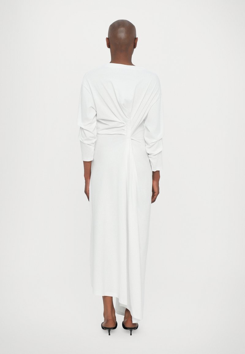 Witte lange mouw jurk met geplooide achterkant, zachte textuur, vloeiende silhouet en asymmetrische zoom. Draagt met zwarte hoge hakken.