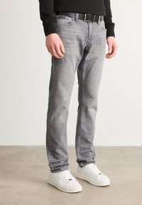 Jeans de mezclilla gris con corte recto, diseño de cinco bolsillos y un cinturón de cuero negro. Combinados con zapatillas blancas sobre un fondo neutro.