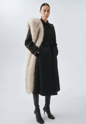 Cappotto nero con un design doppiopetto e uno scialle con accenti di pelliccia. Adottato con una cintura e indossato sopra pantaloni in pelle aderenti e stivali con tacco alto.